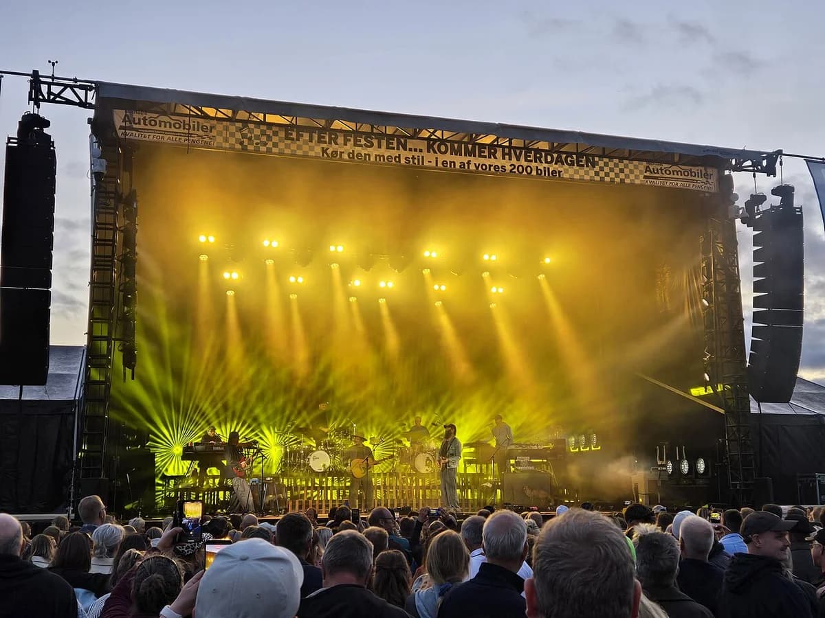 A band performs on a brightly lit outdoor stage in front of a crowd.