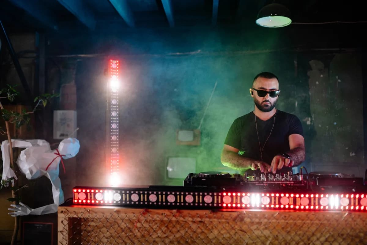 A DJ operating a mixer in a smoky nightclub with colorful lights.
