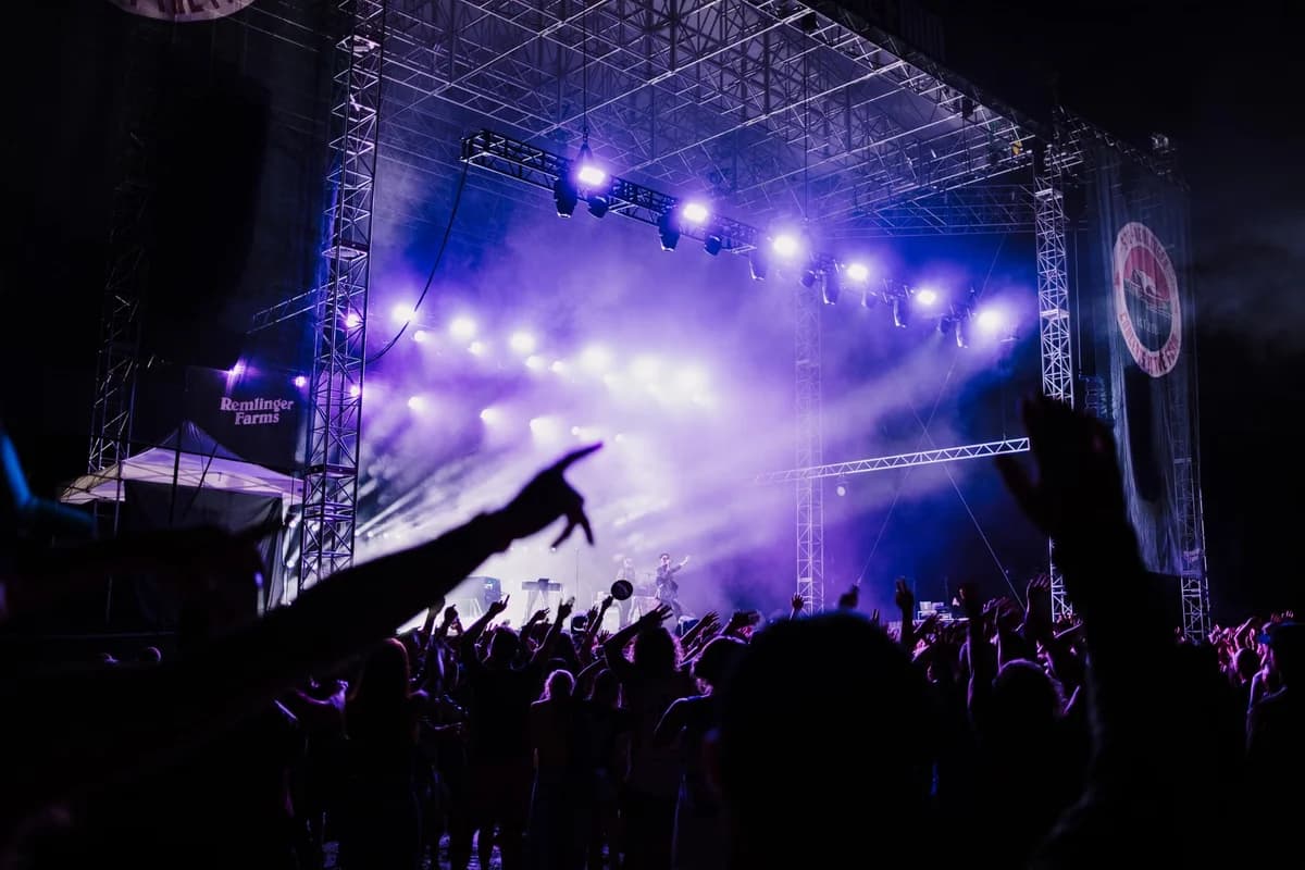 Crowd cheering at an outdoor concert under purple stage lights.