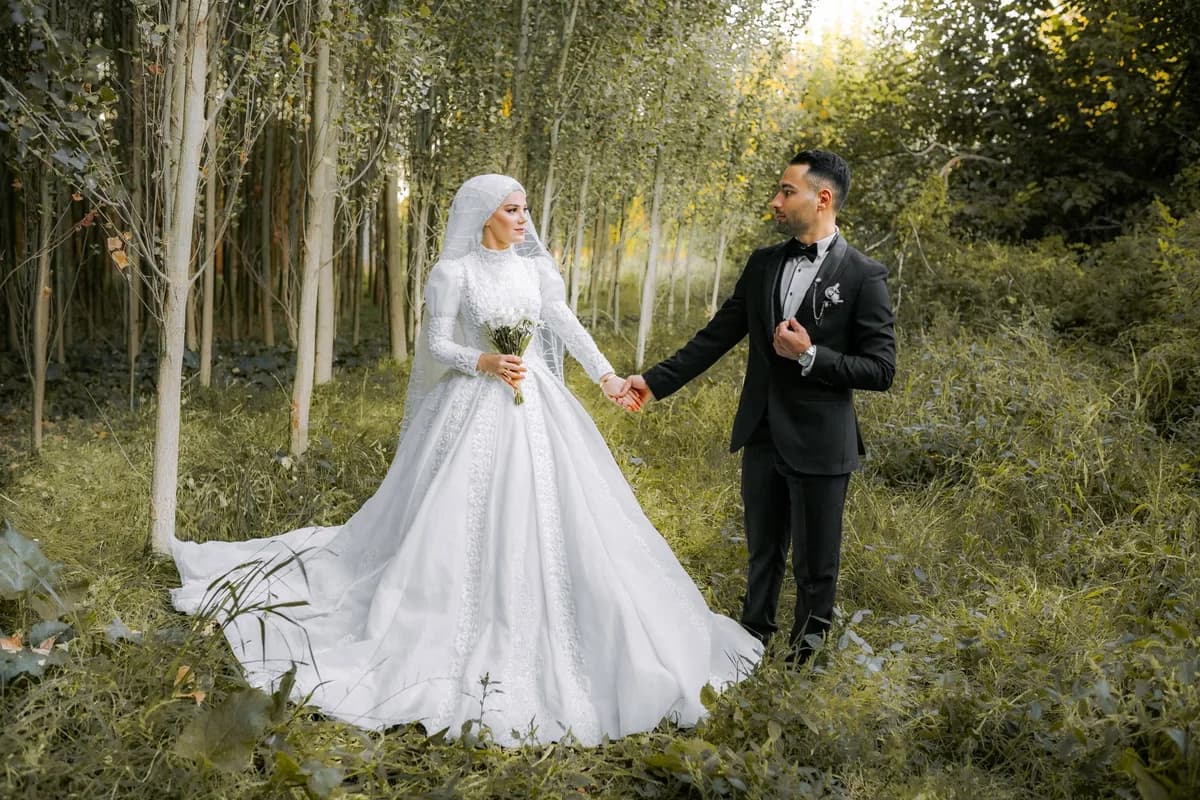 A bride and groom in wedding attire pose hand in hand in a forest clearing.