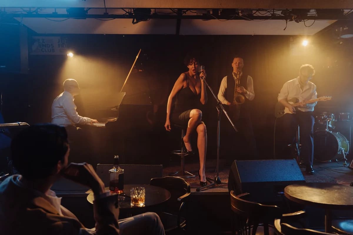 A jazz band performs on a dimly lit nightclub stage with a female singer at center and musicians playing piano, saxophone, and guitar.