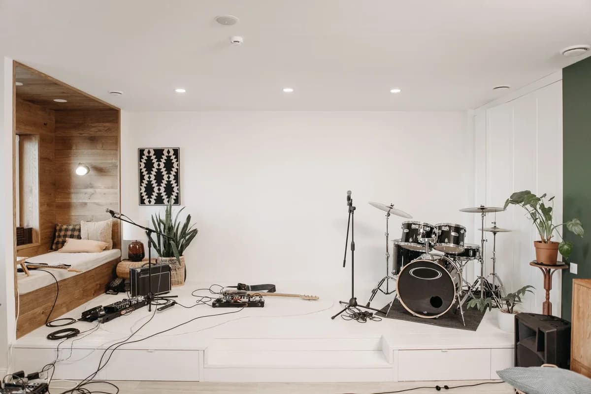 A modern music studio with a drum kit, guitar pedals, a wooden seating nook, and potted plants.