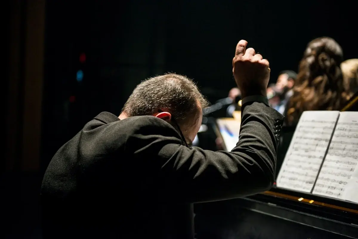 A pianist performs onstage with sheet music in front of him and other musicians blurred behind.