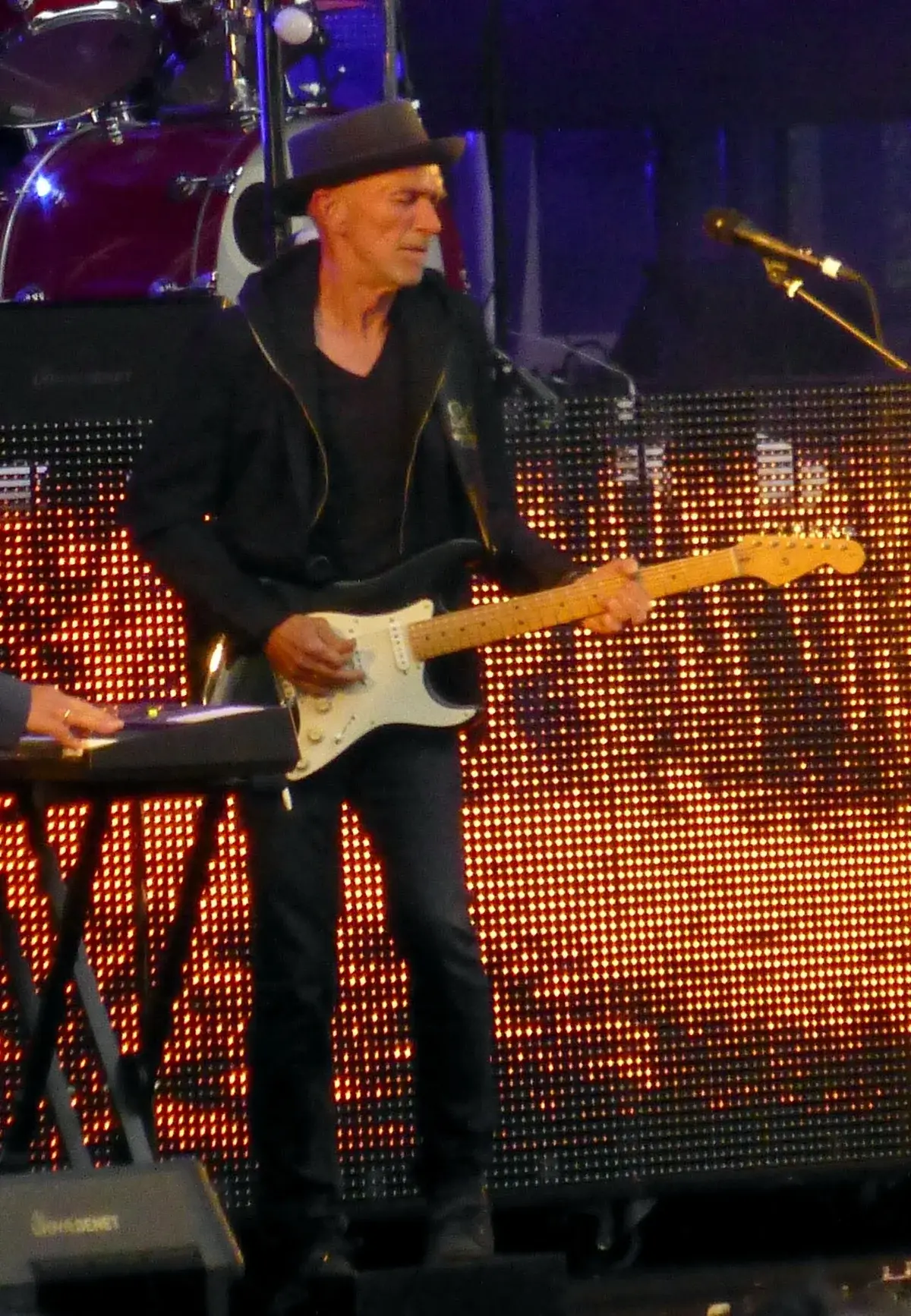 A guitarist in a black hoodie and hat performs onstage in front of a glowing LED screen.