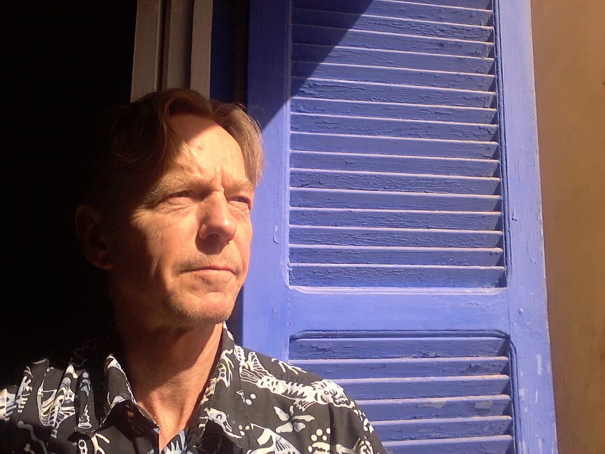 Portrait of a man standing in sunlight beside a blue shuttered window.