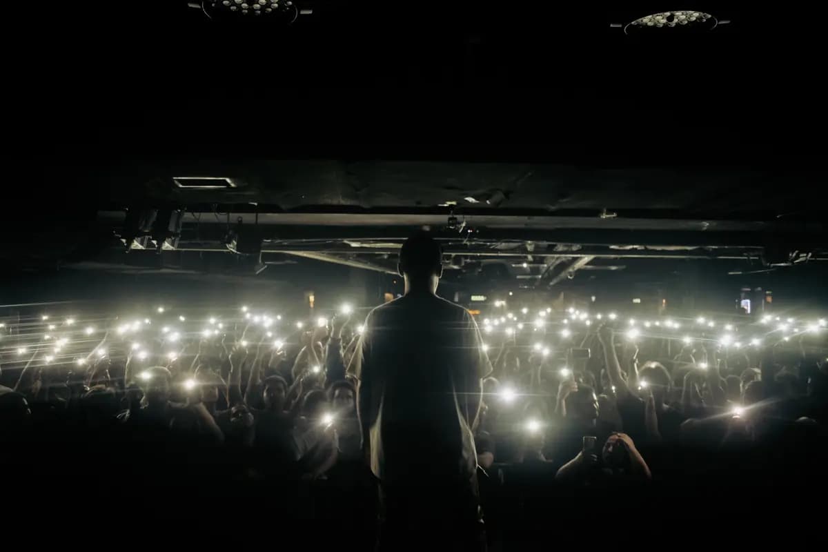 Silhouette of a performer on stage facing an illuminated audience with phone lights at a concert.