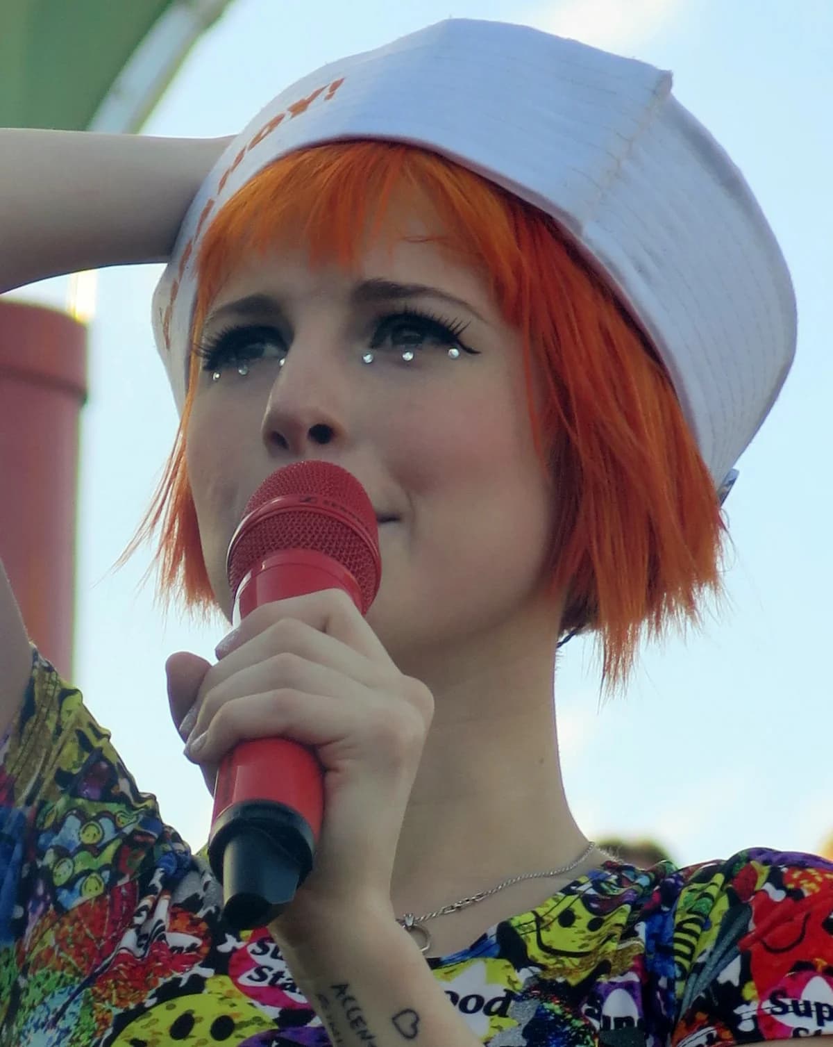 Close-up of Hayley Williams singing into a red microphone while wearing a white hat.