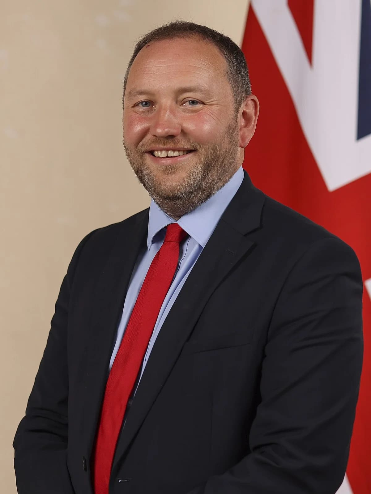 Official portrait of Ian Murray in a suit and red tie, standing in front of a British flag.