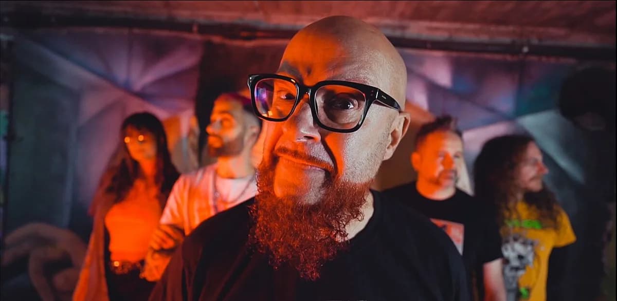 A group of people posing indoors under orange and blue lighting, with a bald man and red beard in the foreground.