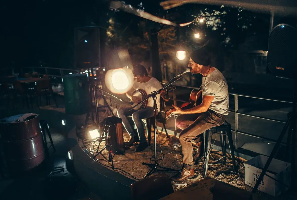 Two musicians performing acoustically on a small outdoor stage at night under warm lights.