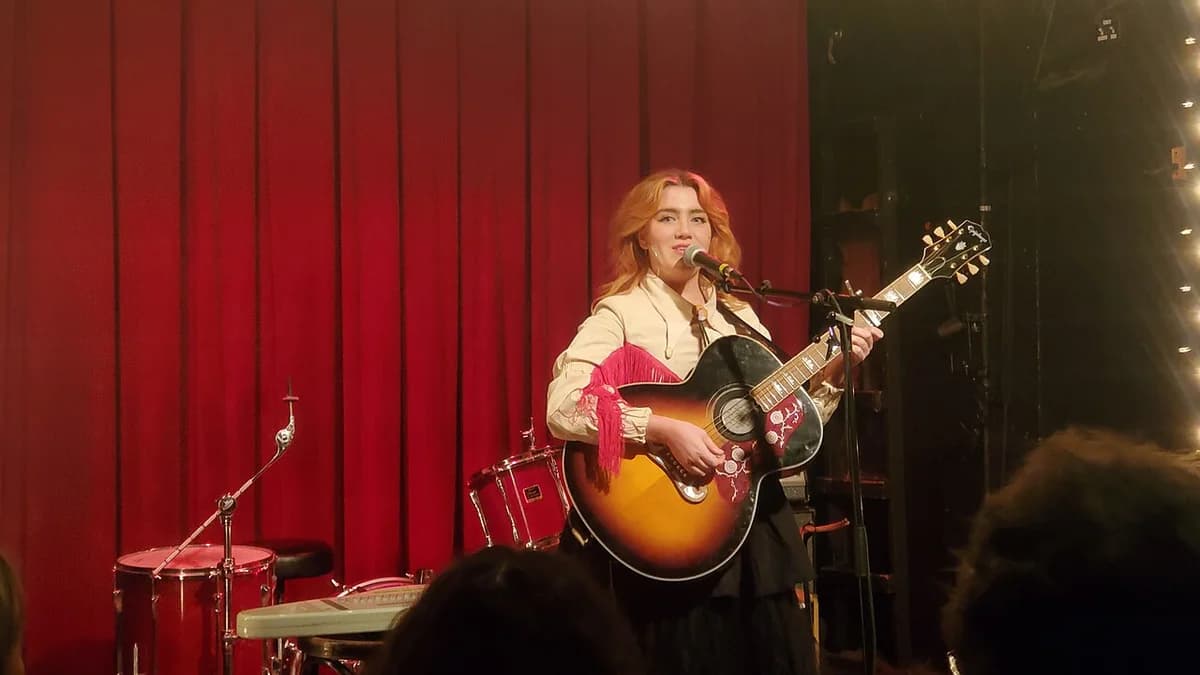 Female musician playing guitar and singing on stage with red curtain background