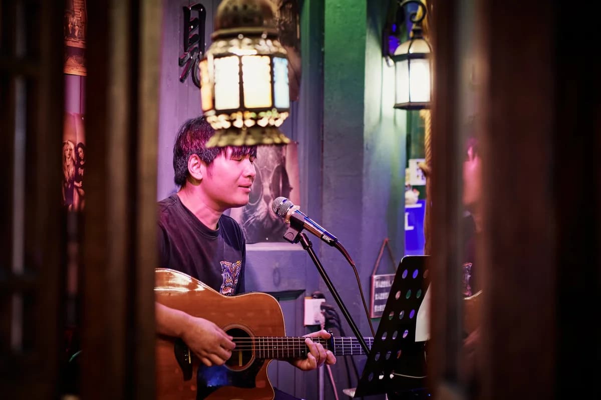 A man sings into a microphone while playing acoustic guitar indoors.