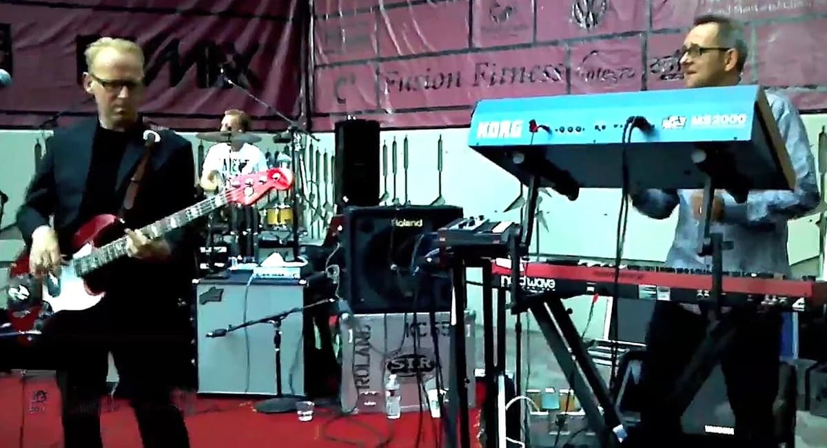 A band performing live on an outdoor stage with a bassist, drummer, and keyboardist.