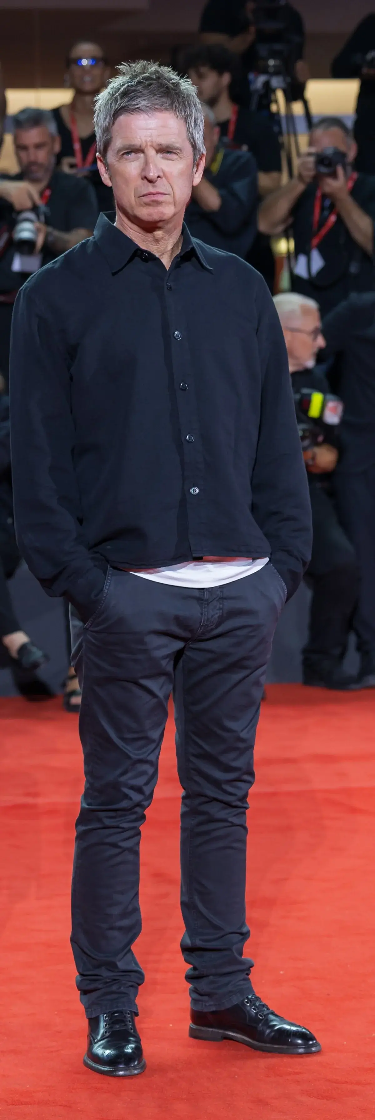 A man poses on a red carpet with photographers behind him.