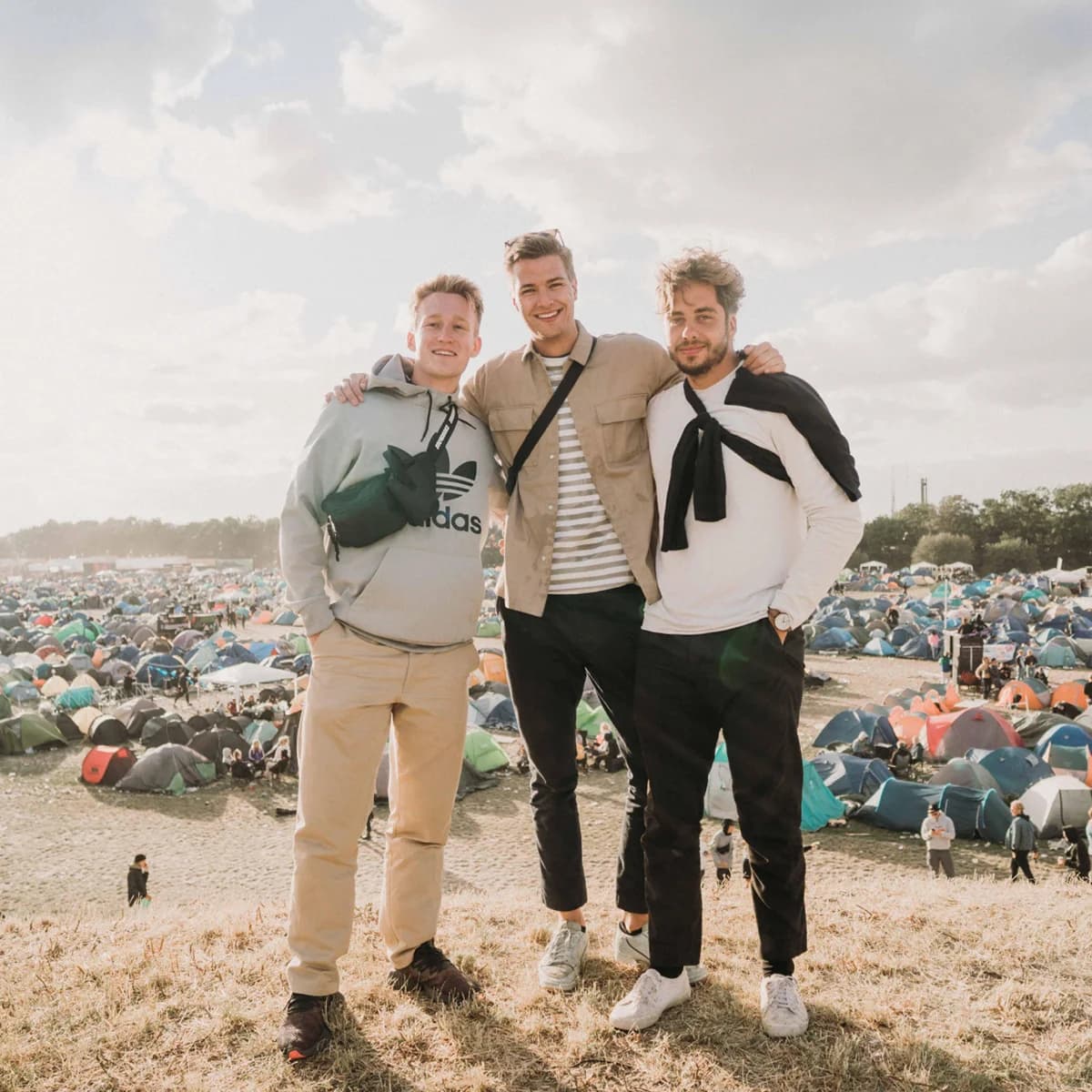 Three men standing together at Roskilde Festival with a large campsite behind them.