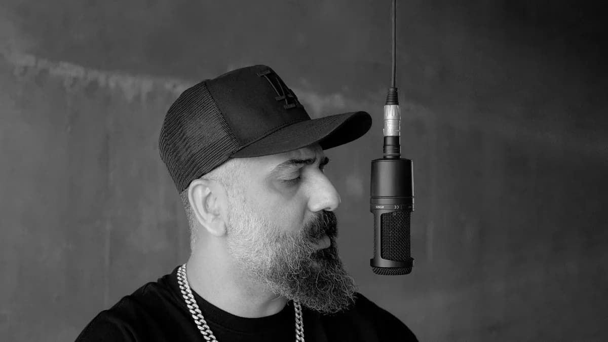 Black-and-white portrait of a bearded rapper wearing a cap beside a microphone.