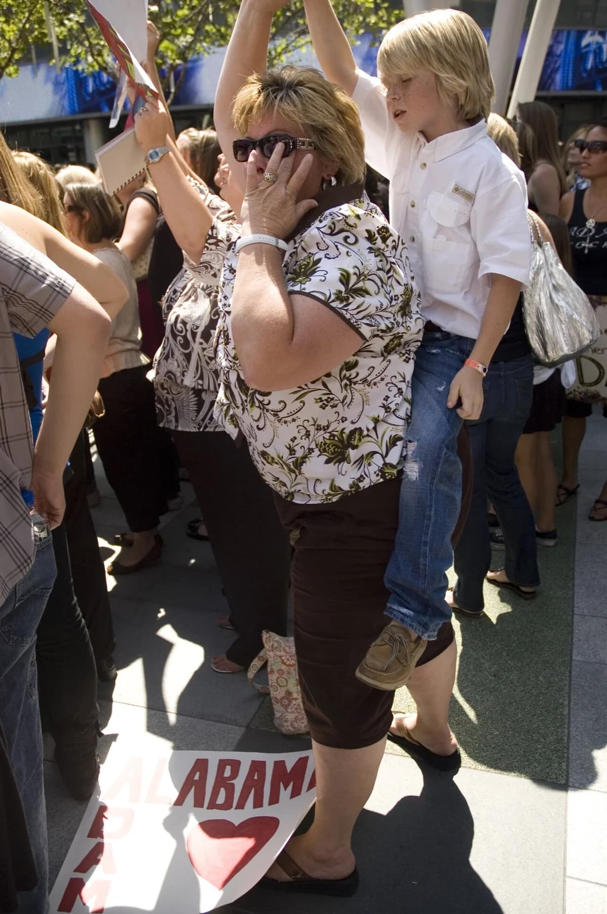 A crowd gathers outdoors, with a woman carrying a boy on her shoulders and signs visible on the ground.