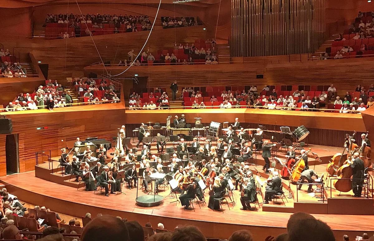 A symphony orchestra performing in a concert hall with an audience seated around the stage.