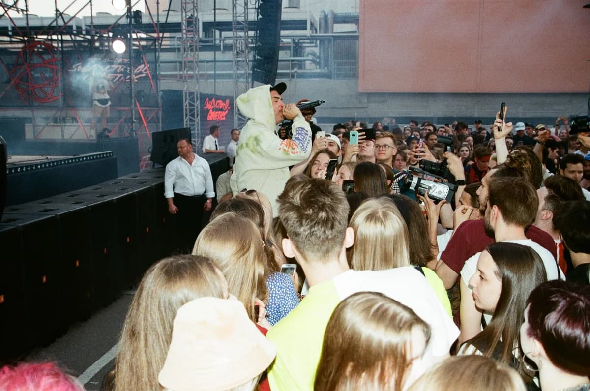 A singer in a white hoodie performs on stage in front of a crowded audience holding up phones.