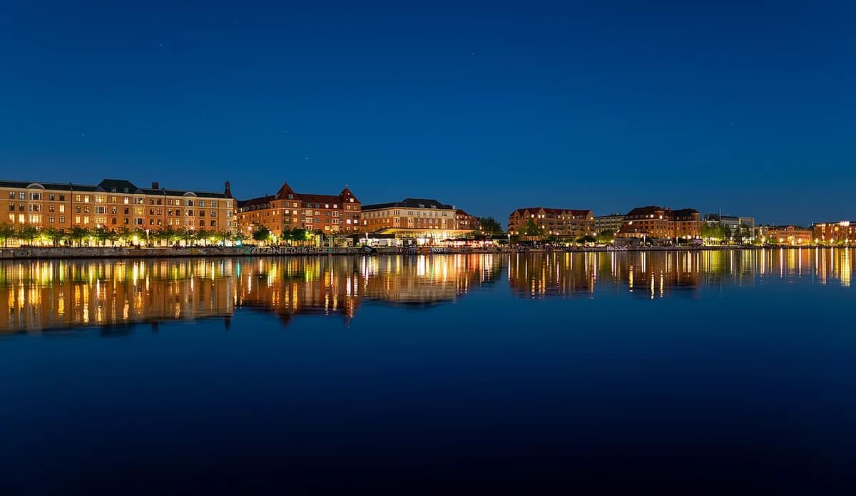 Night view of a canal waterfront with lit buildings reflected in calm water.