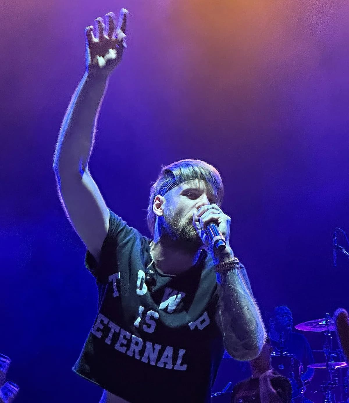 Male rock singer on stage holding microphone with raised hand, drummer in background