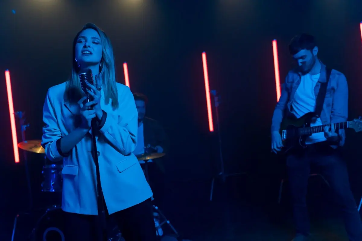 A female singer, guitarist, and drummer perform onstage under blue and red neon lights.