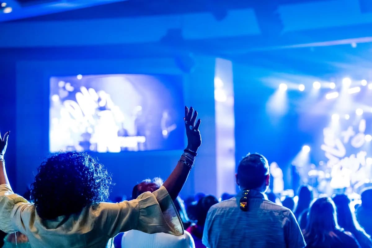 Audience members watch a live concert under blue stage lighting, with one person raising both hands.