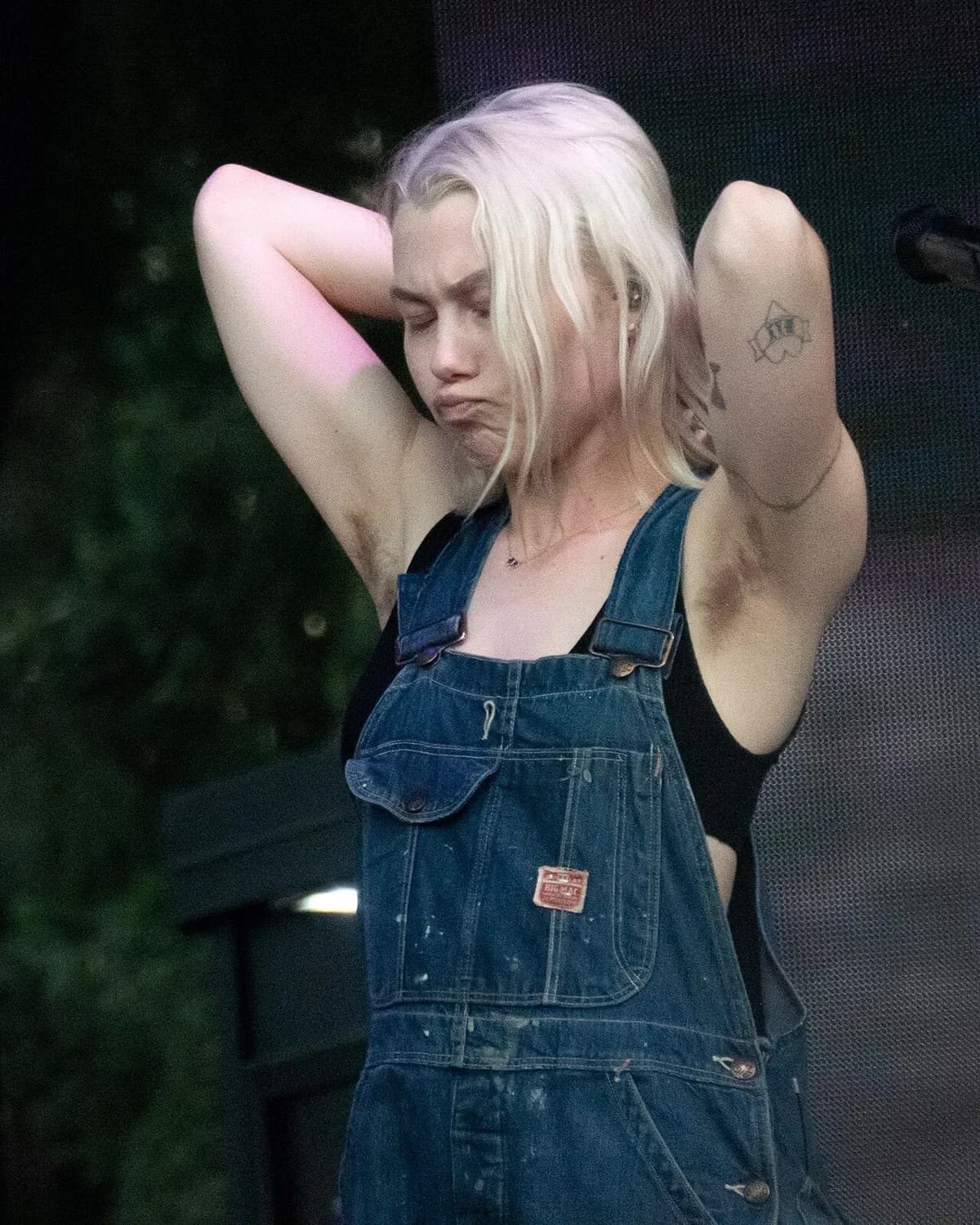 Blonde female musician in denim overalls with hands behind her head on stage