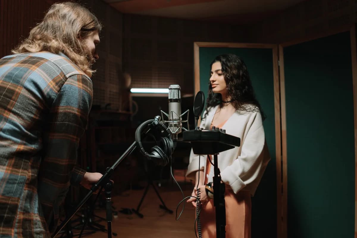 A woman stands at a microphone in a recording studio while a man adjusts the mic stand.