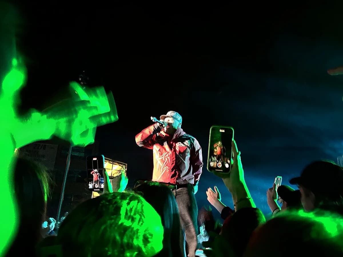A rapper performs on stage under green lights while the audience films with phones.