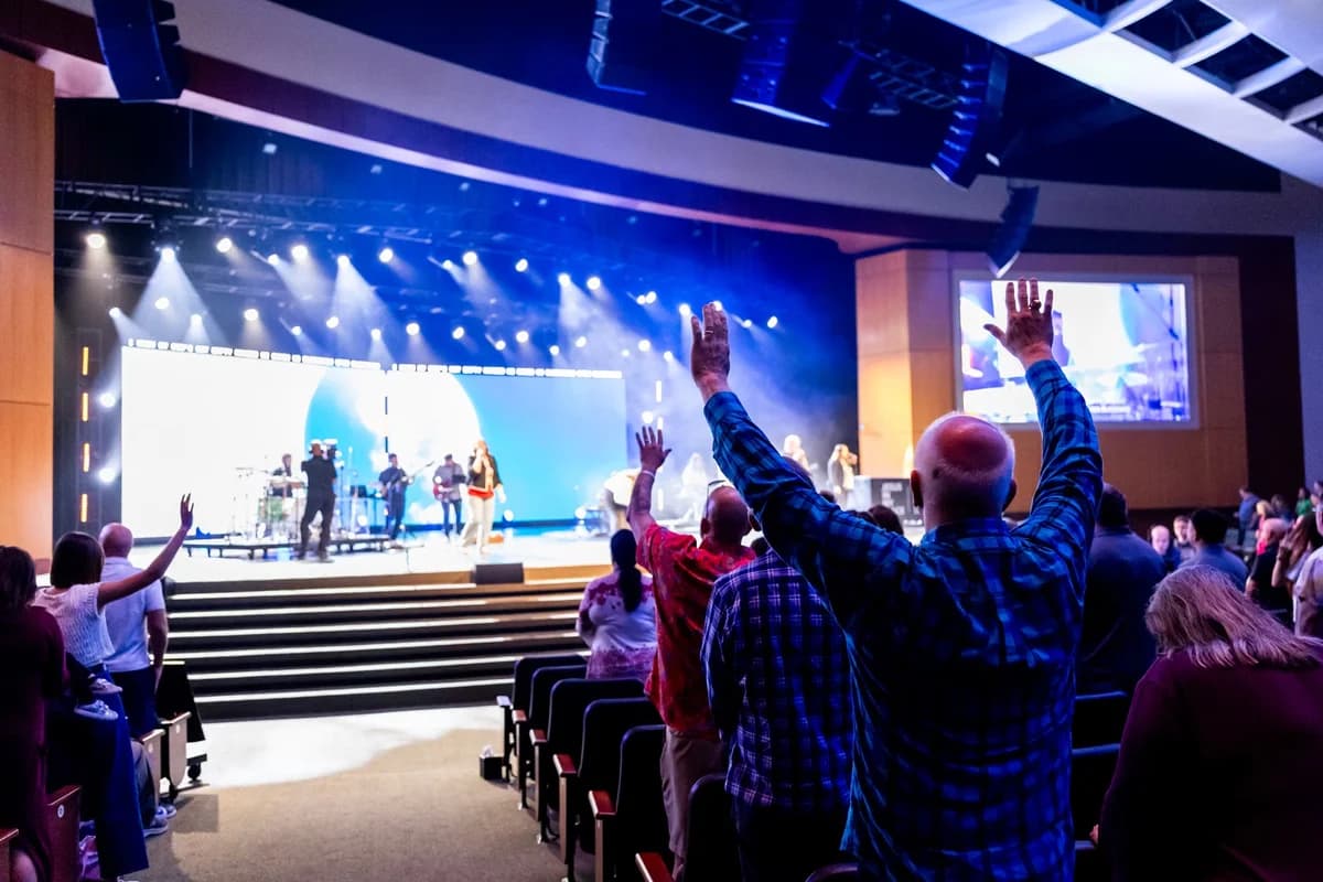 Church audience raising hands during a worship concert with a band on stage.