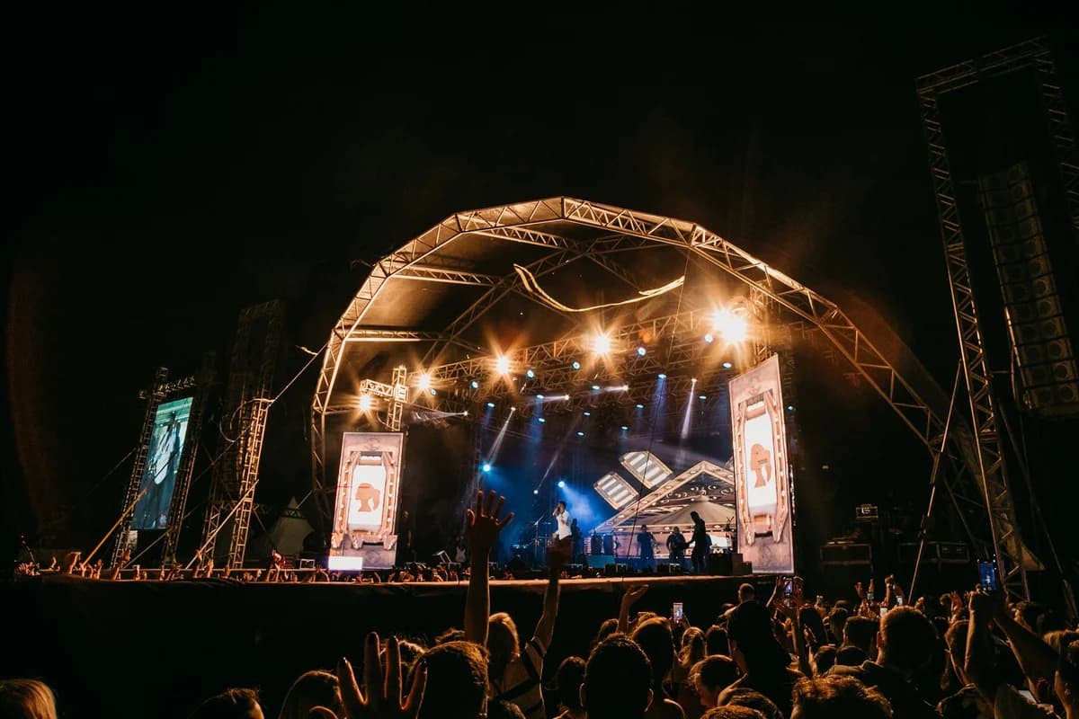Crowd cheering at a nighttime outdoor concert with bright stage lights and large screens.