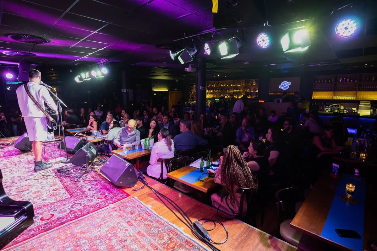A guitarist performs on a small stage in a crowded, dim club with colorful lighting.