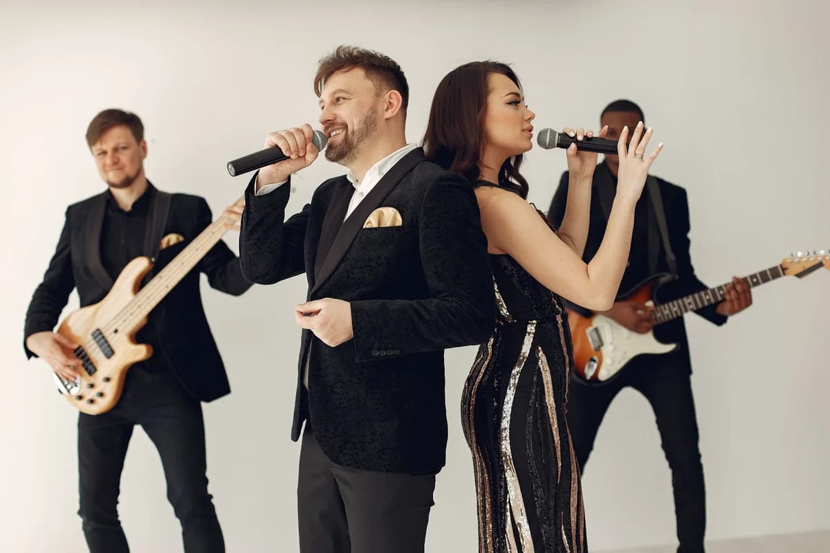 A man and woman sing into microphones while two guitarists perform behind them.